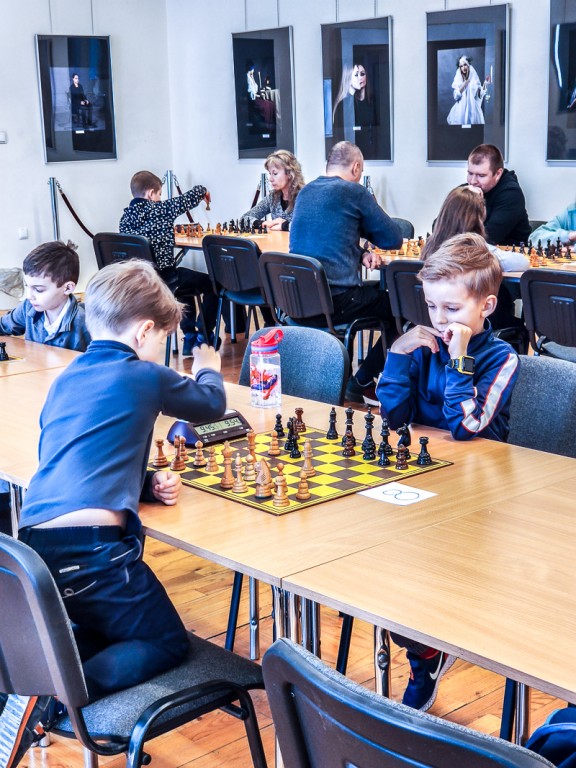 Dwójka dzieci gra w szachy, w tle widać dorosłych grających w szachy