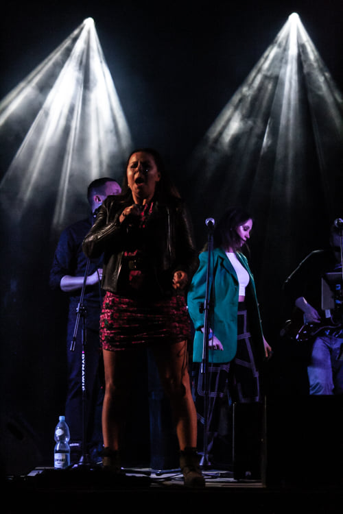 Kobieta w sukience śpiewa na scenie, w tle widać inne osoby