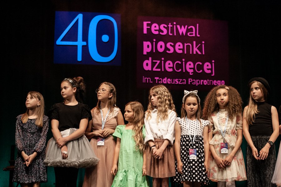 Zdjęcie przedstawia dziewczynki stojące w rzędzie obok siebie, w tle widać logo festiwalu