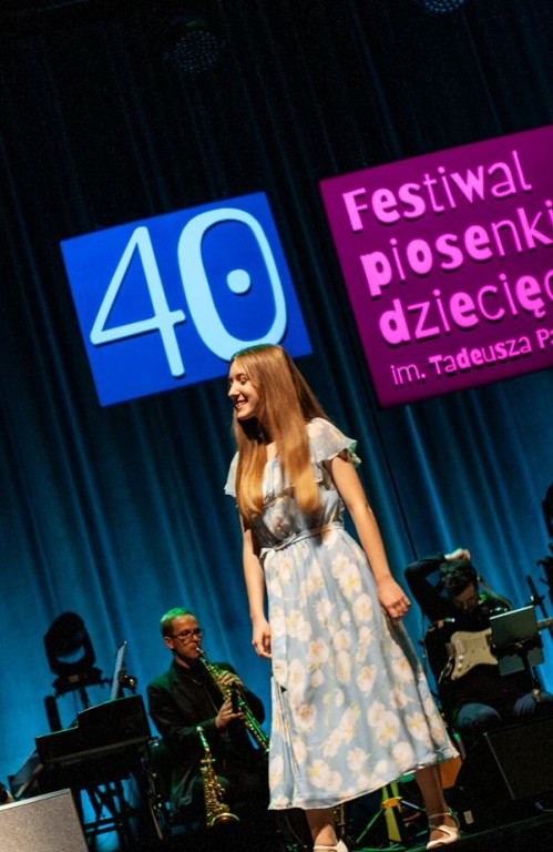Kobieta w długiej sukience występuje na scenie, w tle widać muzyków