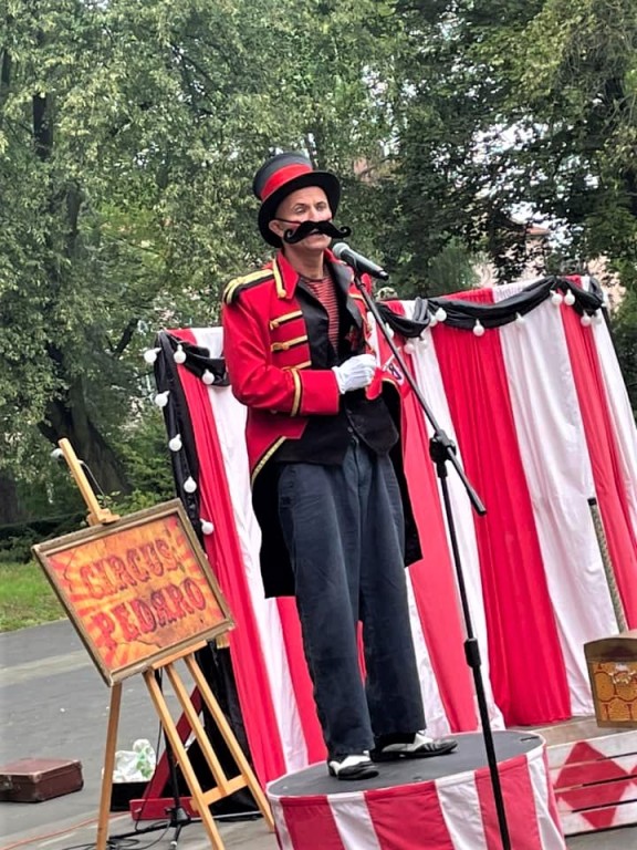 Mężczyzna przebrany za cyrkowca w sztucznych wąsach przemawia na piedestale przed mikrofonem, za nim mała kurtyna w czerwono-białe paski