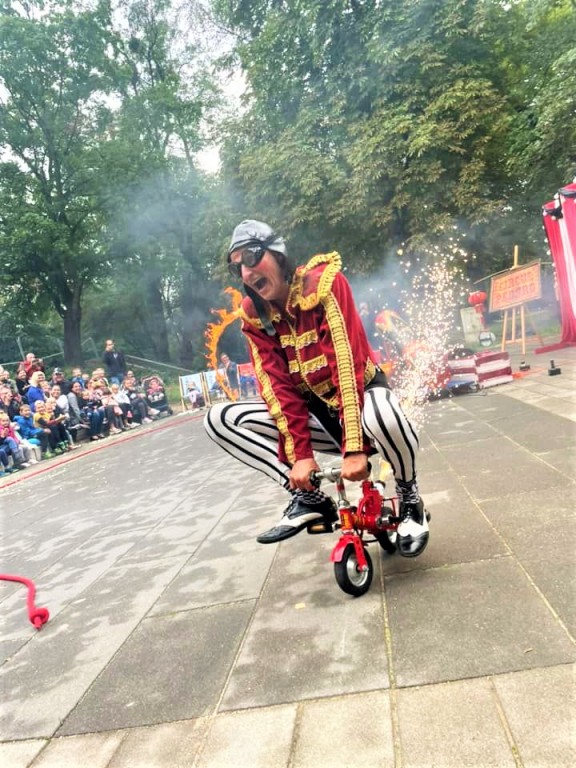 Kobieta przebrana za cyrkowca jadąca na miniaturowym rowerku przed widownią w parku