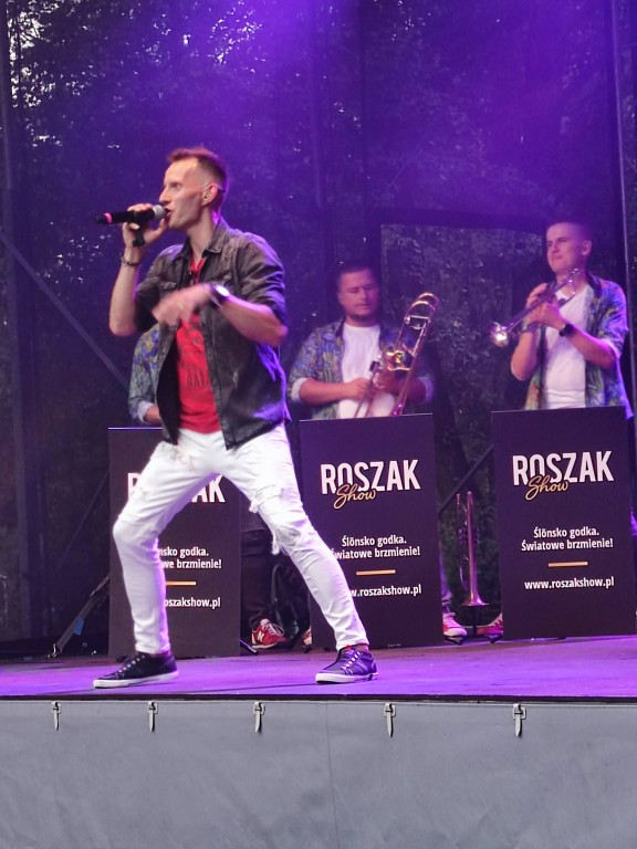 Mężczyzna śpiewający do mikrofonu na scenie, za nim zespół muzyczny, a przed nimi panele podpisane 'ROSZAK'