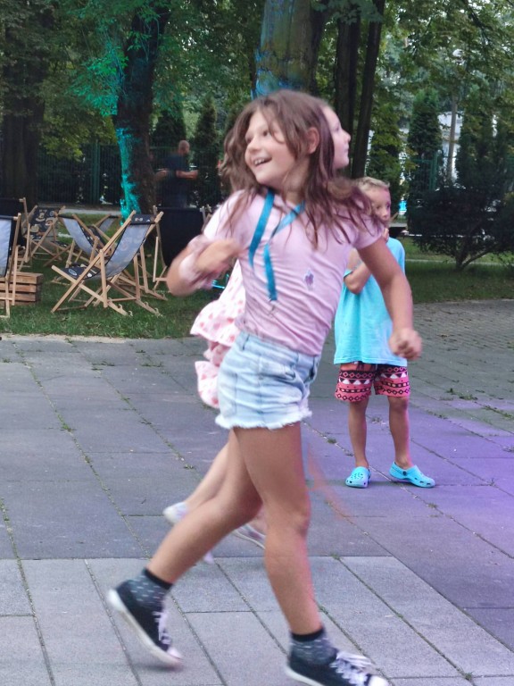 Dziewczynka tańcząca na chodniku w parku, na nią inne dzieci