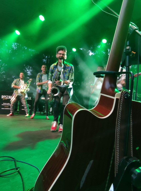 Fragment gitary postawionej na stojaku, za nią zespół muzyczny na scenie oświetlonej w zielonych barwach