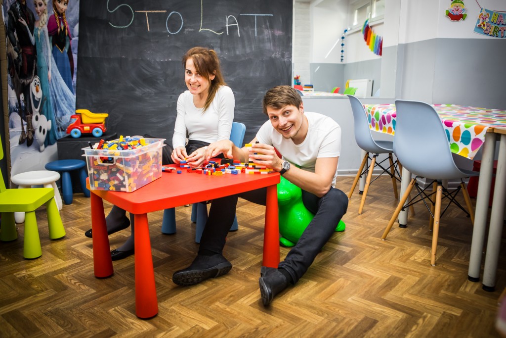 Kobieta z mężczyzną siedzą obok stoliczka dla dzieci, za nimi na tablicy napis 'STO LAT'