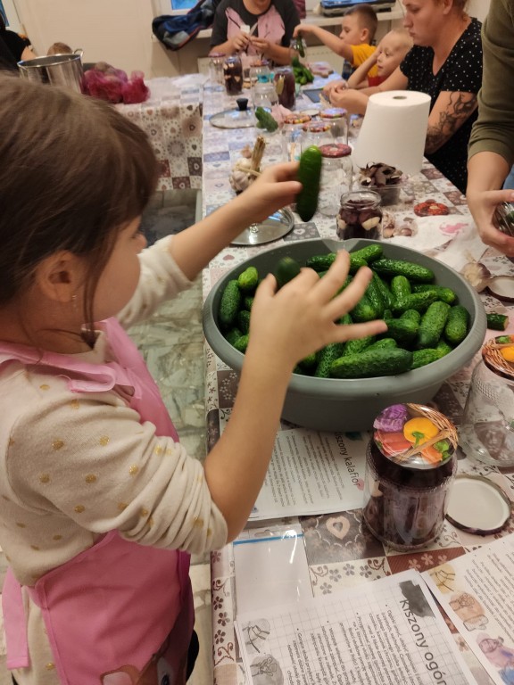 Dziewczynka podnosząca ogórki z miski na stole, przy niej dzieci z opiekunami przygotowują jedzenie.