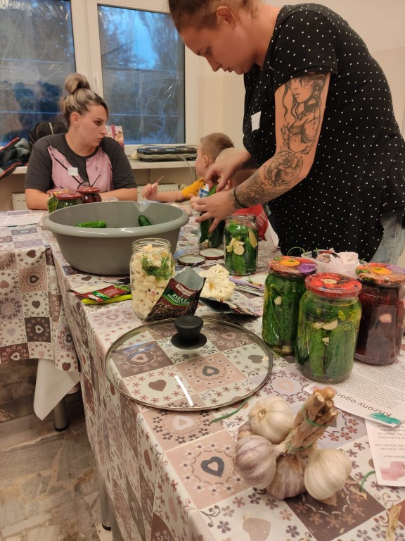 Kobieta przy długim stole krojąca warzywa, za nią inne kobieta rozmawiająca z dzieckiem, a przed słoiki z przetworami oraz czosnek
