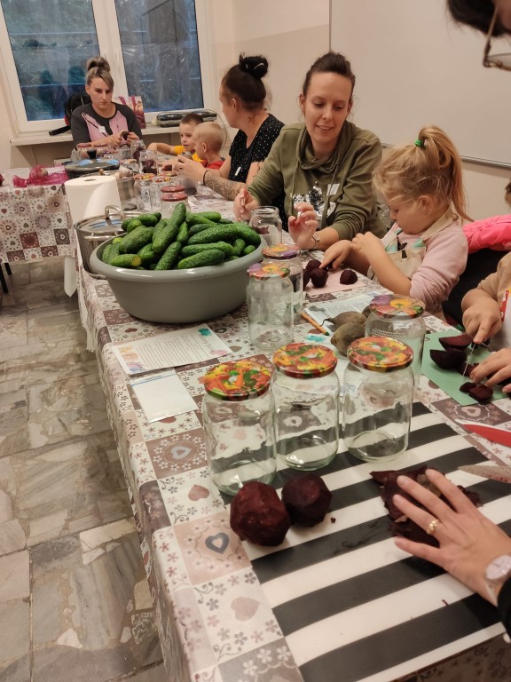 Opiekunki z dziećmi przygotowujące jedzenie przy długim stole, na którym wyłożona jest masa warzyw i naczyń.