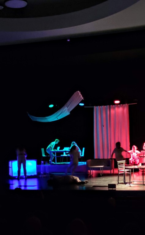 Aktorzy występują na scenie, wokół nich znajdują się stoły z krzesłami i kanapa