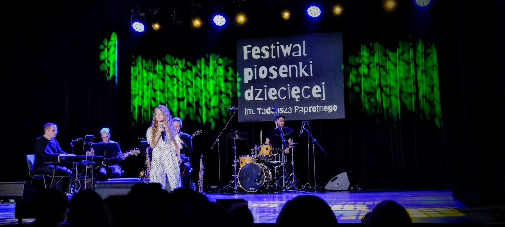 Dziewczynka w białej sukience śpiewa na scenie, w tle widać muzyków