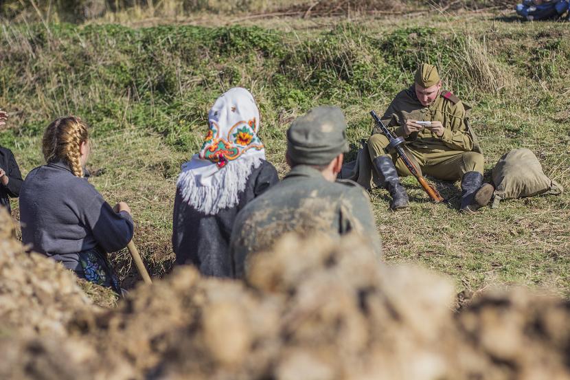 Ludzie siedzący na trawie przed również siedzącym żołnierzem z bronią