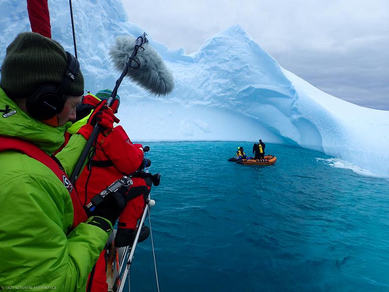 Dwie osoby stojące nad wodą, a w oddali ludzie na łódce, za nimi arktyczna góra lodowa