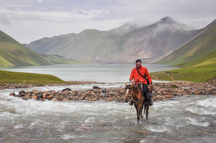 Osoba w czerwonej kurtce jadąca na koniu przez potok na tle gór