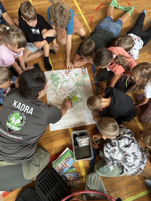 Dzieci wraz z panią przewodnik siedzą wokół mapy. Przewodniczka tłumaczy jak zorientować mapę.