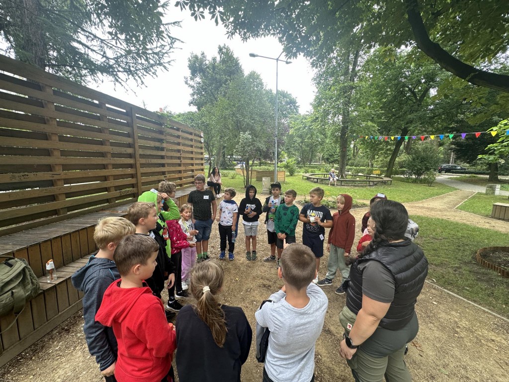 Dzieci stoją w kole i słuchają przewodniczki. Po prawej, zaraz za grupą, widać drewniany płot. Z lewej strony rozciąga się park.