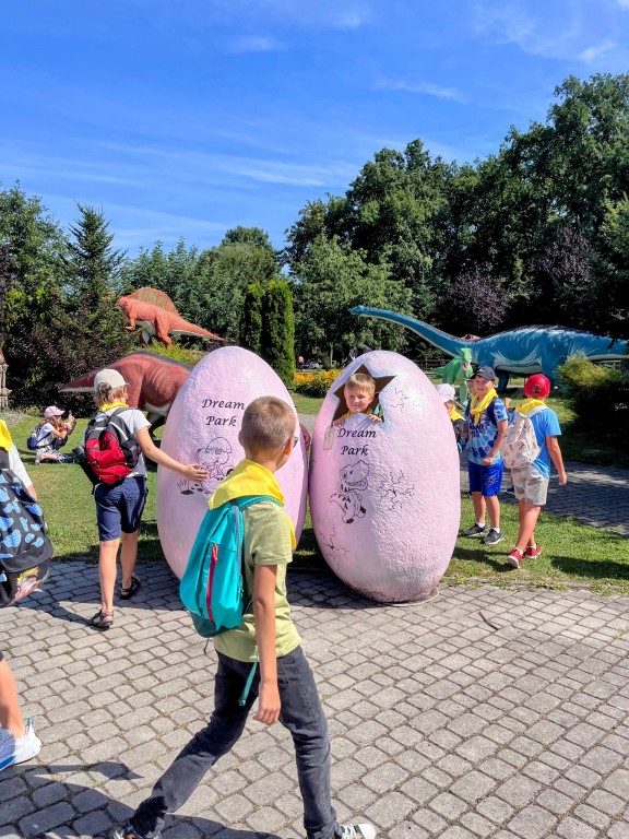 Dzieci pozują do zdjęcia z dużymi jajami dinozaurów. W tle widać cztery kolorowe dinozaury.