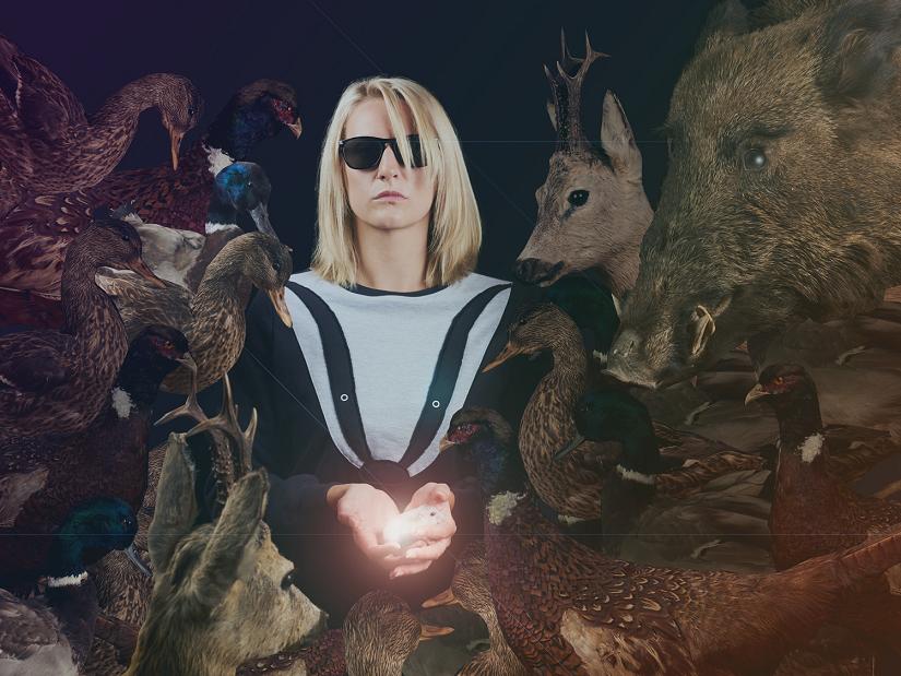 Kobieta w okularach przeciwsłonecznych, w jej dłoniach ukryty świecący przedmiot, grupa wypchanych zwierząt jest skierowana w stronę światła.