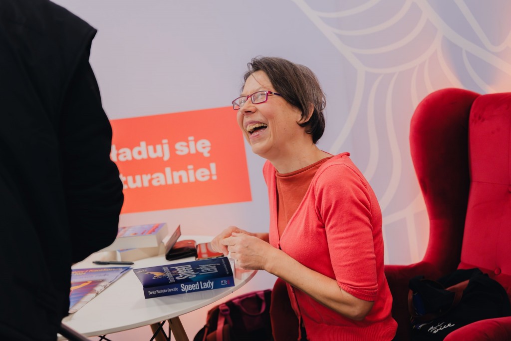 kobieta siedzi na fotelu, jest rozbawiona, patrzy w górę na swojego rozmówcę