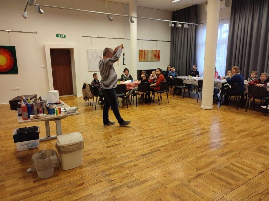 w sali, mężczyzna prezentuje marionetkę, osoby przy stolikach patrzą na niego