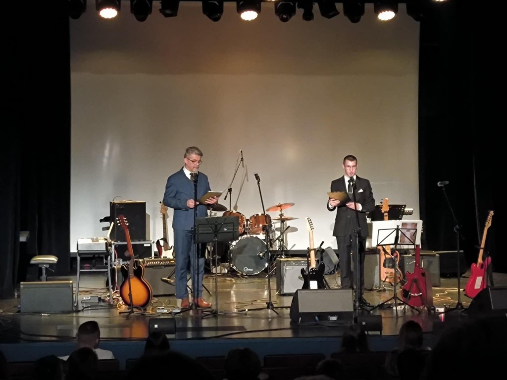 dwóch mężczyzn w garniturach na scenie, w tle ekran i sprzęt muzyczny