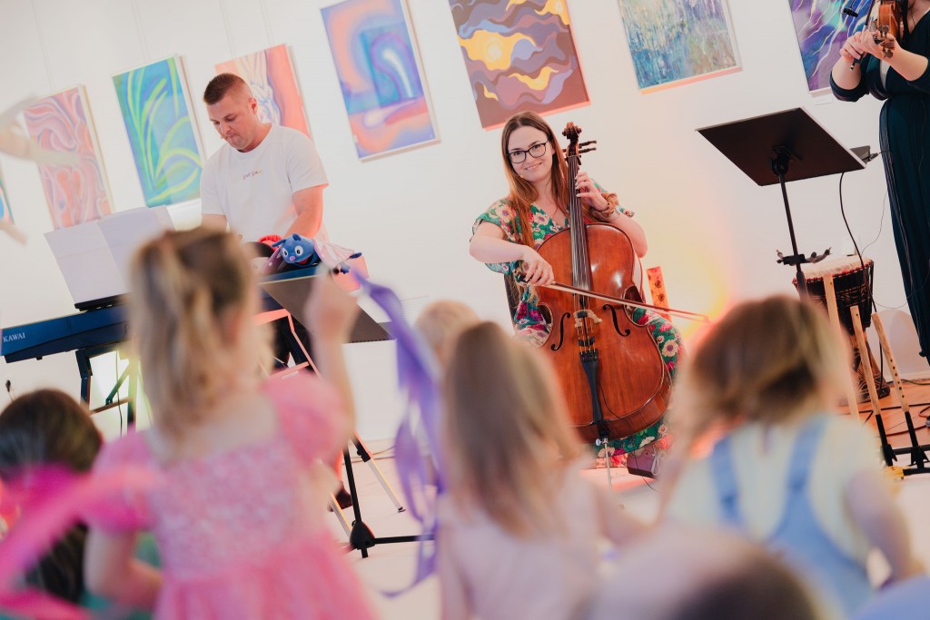 Zespół muzyczny gra na instrumentach dla małych dzieci, występują w jasnym pomieszczeniu, na ścianach wiszą kolorowe obrazy.