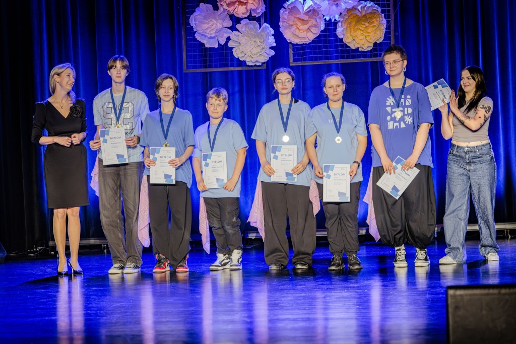 Mała grupa młodzieży w niebieskich strojach stoi na scenie z dyplomami i medalami w rękach.