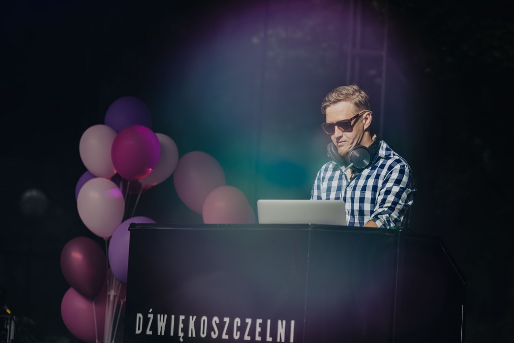DJ stoi na ciemnej scenie z reflektorem, przy rozstawionym sprzęcie i słuchawkami na głowie.