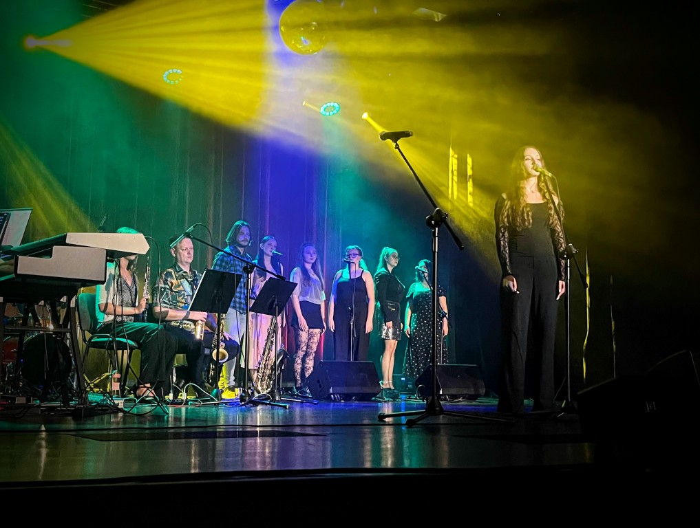 Zespół muzyczny występuje na kolorowo oświetlonej scenie; grają instrumentaliści, śpiewa wokalistka i towarzyszy jej chórek.