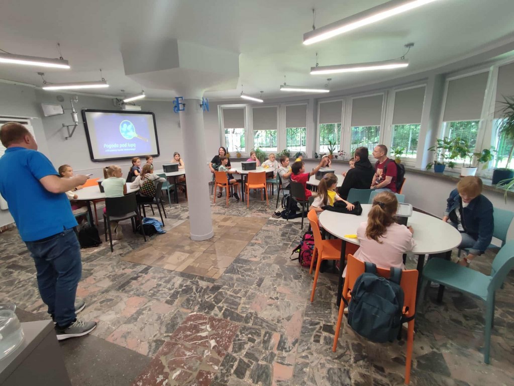 Dzieci siedzą przy stolikach i słuchają Przewodnika.