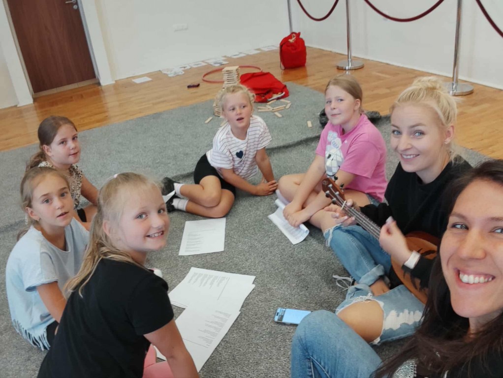 Dzieci siedzą w kółku z opiekunkami i słuchają jak jedna z nich gra na ukulele.