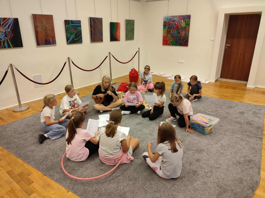 Dzieci siedzą w kółku z opiekunką i razem śpiewają.