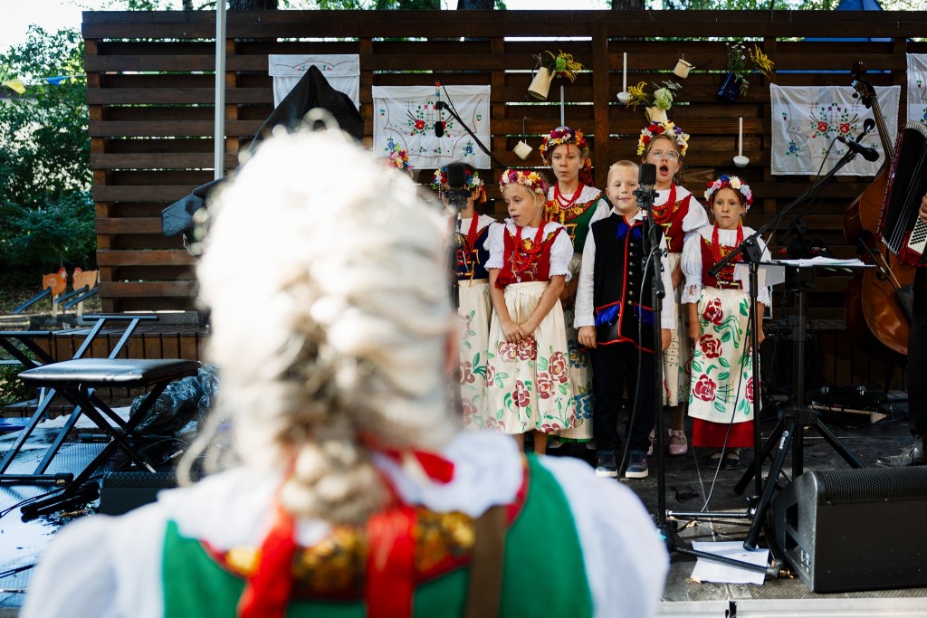 Widok na scenę na której występują dzieci w strojach ludowych, patrzymy zza pleców kobiety. Widać jej rozmazaną głowę.
