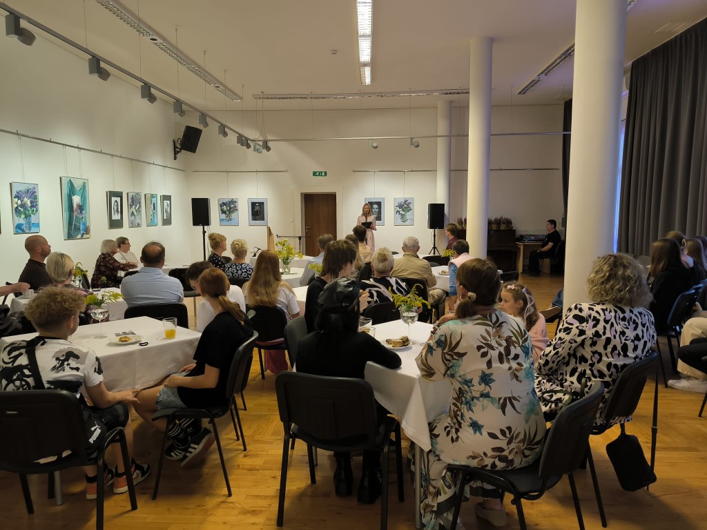 3.	Widok na salę, w tle kobieta mówi do mikrofonu, ludzie siedzą przy stolikach (ustawienie kawiarniane).