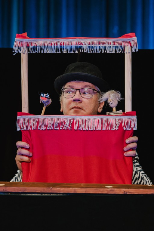 mężczyzna zerka z małej, miniaturowej sceny, trzyma pacynki.