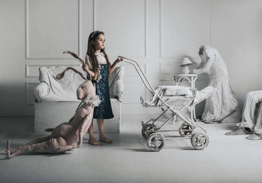 Dziewczynka w białym pomieszczeniu trzymająca dużego pluszowego zając oraz prowadząca biały wózek dla dziecka