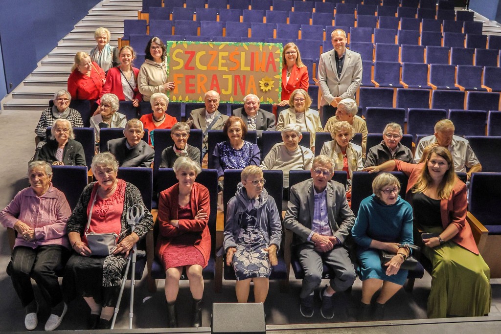 Na widowni siedzą seniorzy na pierwszych trzech rzędach foteli,w czwartym rzędzie dwie panie trzymają plakat kolorowy z napisem SZCZĘŚLIWA FERAJNA.