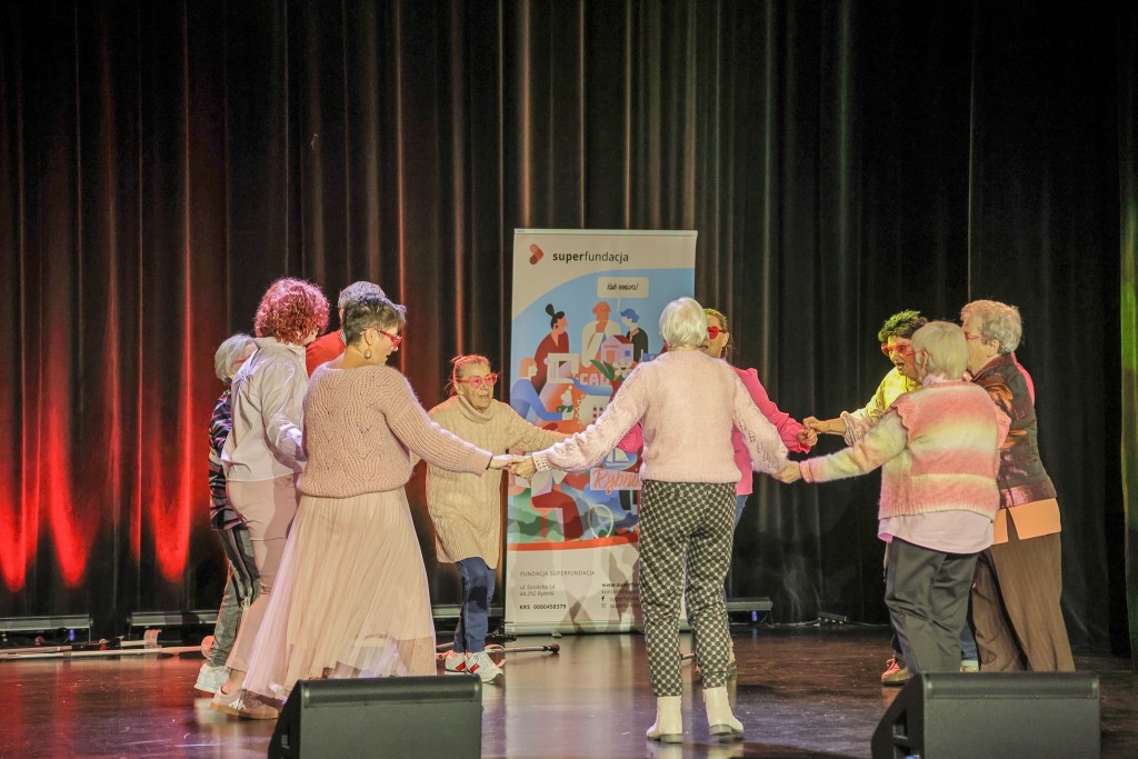 Panie seniorki stoją na scenie trzymają się za ręce w kołeczku, są ubrane na różowo mają także różowe okularki w kształcie serc koło nich stoi plakat z super fundacji.