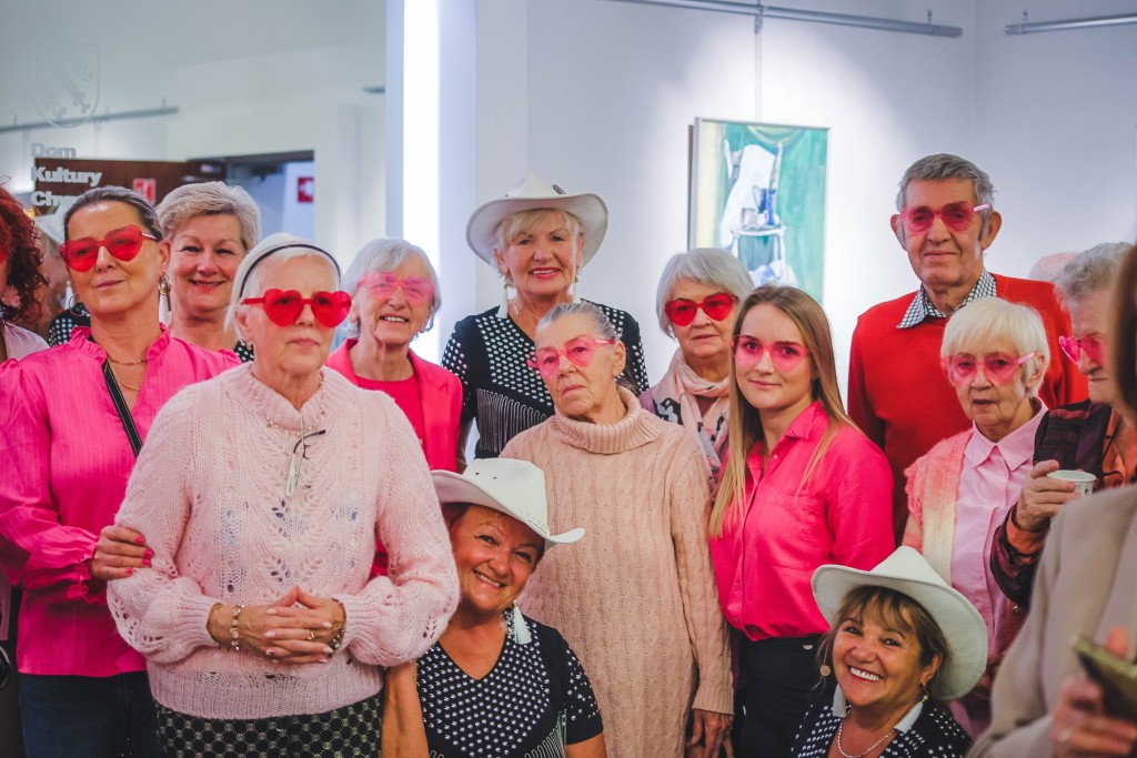 seniorzy w ustawieni po występie do zdjęcia maja różowe ciuchy i okularki w serca a niektóre panie maja kowbojskie białe kapelusze i czarne stroje.