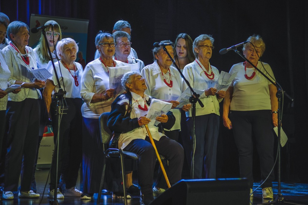 Seniorzy stoją na scenie , a jedna pani siedzi na krześle, przed nimi stoją trzy mikrofony, na szyi seniorki mają czerwone korale i są ubrane w białe koszule ,bluzki  .W rekach trzymają teksty piosenek z których śpiewają