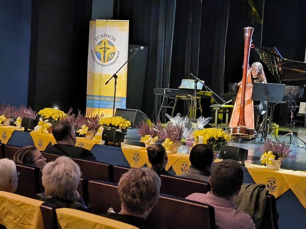 Na zdjęciu widać scenę ozdobioną żółtymi kwiatami i dekoracjami z logotypem Fundacji „Dzieło Nowego Tysiąclecia”. Na scenie siedzi harfistka grająca na dużej harfie, a obok niej znajdują się instrumenty muzyczne — pianino i keyboard. W tle widoczny jest b
