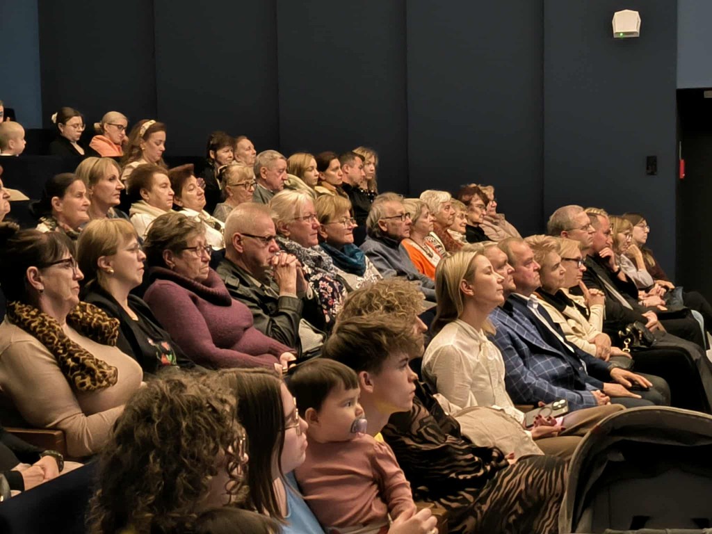 Na zdjęciu widać publiczność siedzącą w sali widowiskowej. Ludzie w różnym wieku – od dzieci po osoby starsze – uważnie obserwują scenę, skupieni na odbywającym się wydarzeniu. Wszyscy siedzą w rzędach na widowni.