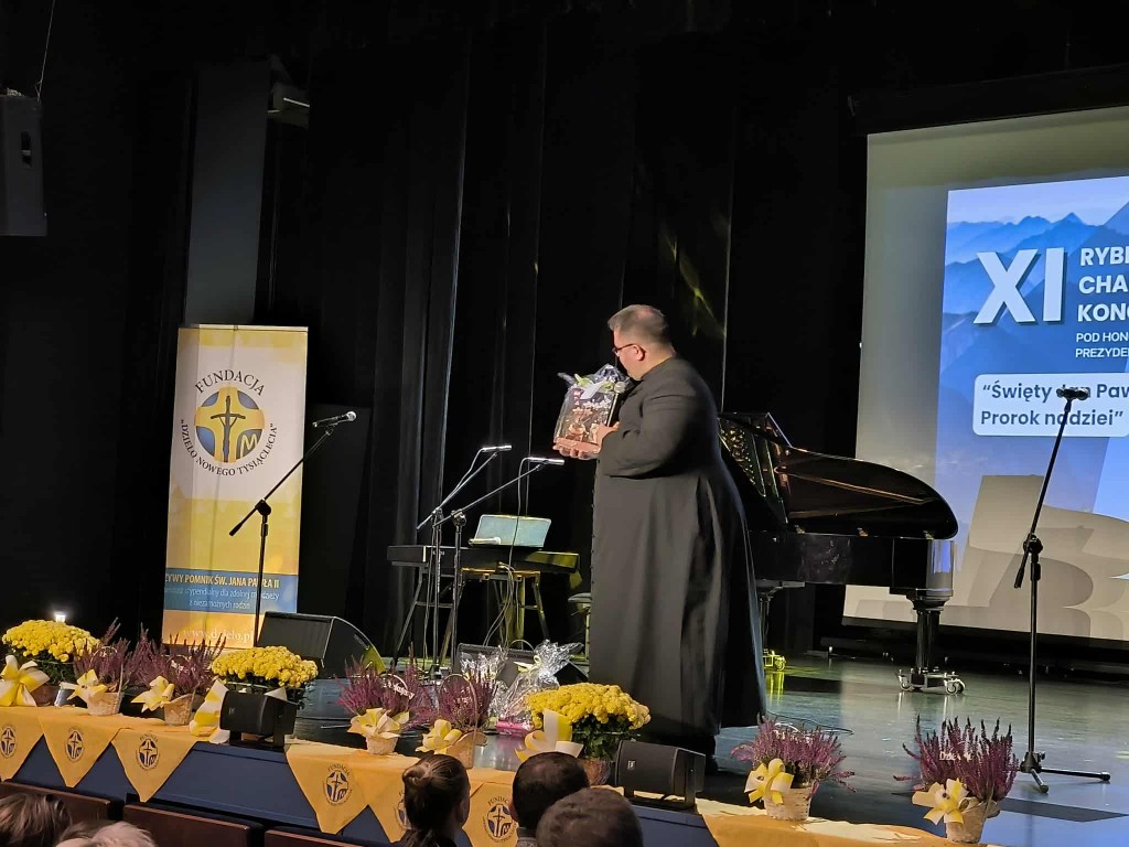 Na zdjęciu widać scenę udekorowaną żółtymi kwiatami i materiałami z logotypem Fundacji „Dzieło Nowego Tysiąclecia”. Na scenie stoi duchowny w czarnej sutannie, trzymający w rękach kosz z upominkiem. W tle widoczny jest ekran z napisem „XI Rybnicki Charyta