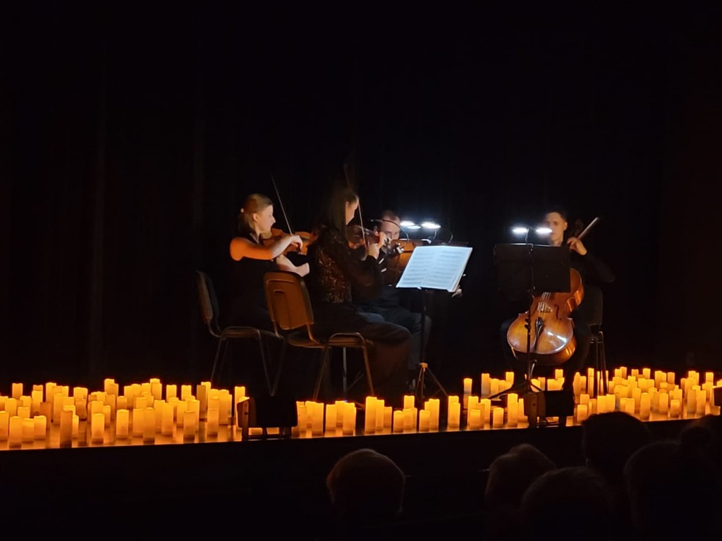 Widok na scene z lewej strony przybliżone zdjęcie.Po środku siedzą muzycy ,cztery osoby;dwie panie i pan grają na skrzypcach , a jeden pan na kontrabasie,przed soba mają nuty podświetlone, postawione na statywach ,wokół nich są świece o pomarańczowym blas