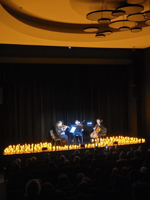Widok na scene z lewej strony ,oddalony.Po środku siedzą muzycy ,cztery osoby;dwie panie i pan grają na skrzypcach , a jeden pan na kontrabasie,przed soba mają nuty podświetlone, postawione na statywach ,wokół nich są świece o pomarańczowym blasku ,które 