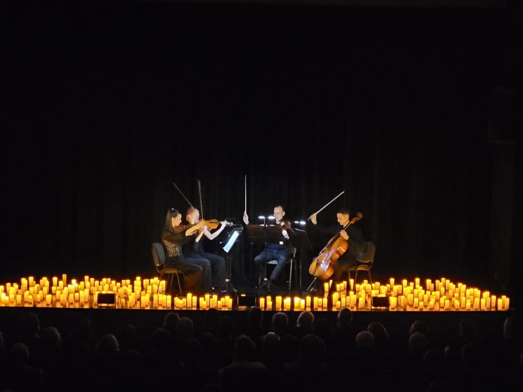 Widok na wprost sceny ,przybliżone zdjęcie.Po środku siedzą muzycy ,cztery osoby;dwie panie i pan grają na skrzypcach , a jeden pan na kontrabasie,przed soba mają nuty podświetlone, postawione na statywach ,wokół nich są świece o pomarańczowym blasku ,któ