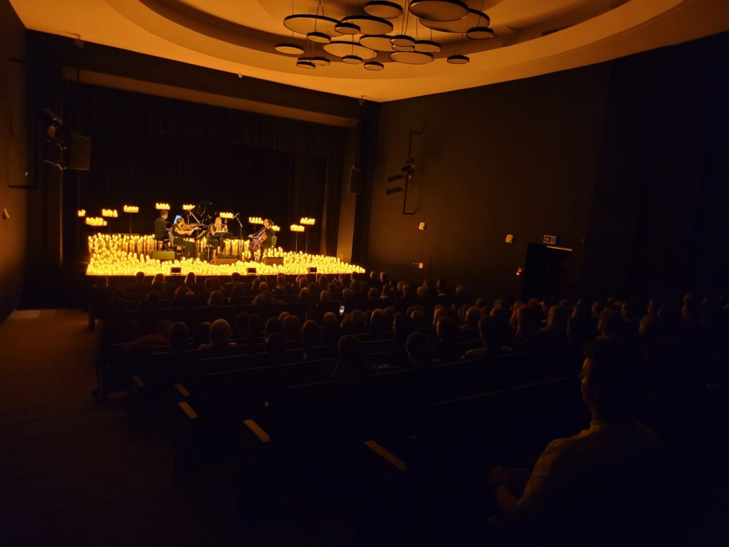 Widok z ostatnich rzędów po lewej stronie. Po środku sceny siedzą cztery osoby;dwie kobiety grają na skrzypcach , jeden mężczyzna gra na kontrabasie, a drugi na pianinie za nimi, przed sobą mają nuty, postawione na statywach ,wokół nich są świece o pomara
