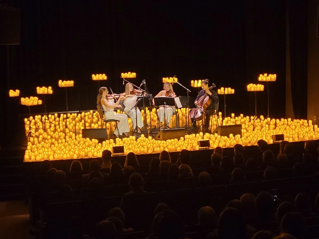 Przybliżony widok czterech osób,siedzą po środku sceny ;trzy kobiety grają na skrzypcach , jeden mężczyzna gra na kontrabasie, przed sobą mają nuty, postawione na statywach ,wokół nich są świece o pomarańczowo-żółtym blasku ,które robią przytulny klimat. 