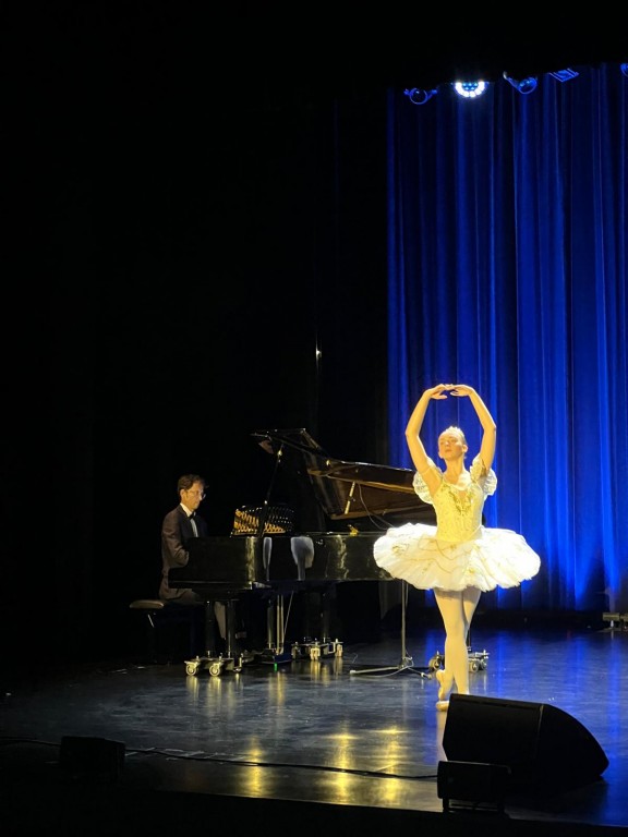 Widok na scene z prawej strony. Baletnica stoi na palcach w eleganckiej pozie, z uniesionymi nad głowę ramionami tworzącymi koło, w klasycznym, białym kostiumie scenicznym i tiarą. Za baletnicą widać pianistę grającego przy fortepianie, a całość oświetlon