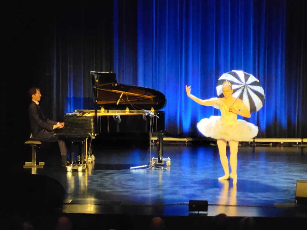 Baletnicę tańczącą na scenie stoi obok pianisty grającego na fortepianie. Tancerka ubrana jest w jasny, klasyczny kostium baletowy i tiarę, a w ręku trzyma czarno-białą otwartą parasolkę jakby padał deszcz. Stoi na jednej nodze, drugą lekko unosi przed so